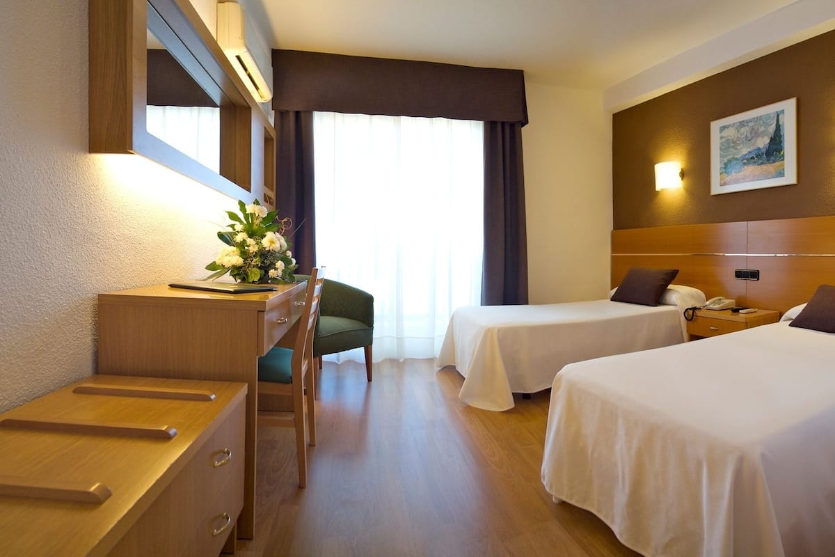 A hotel room with two beds, a desk, a chair, and a desk lamp.