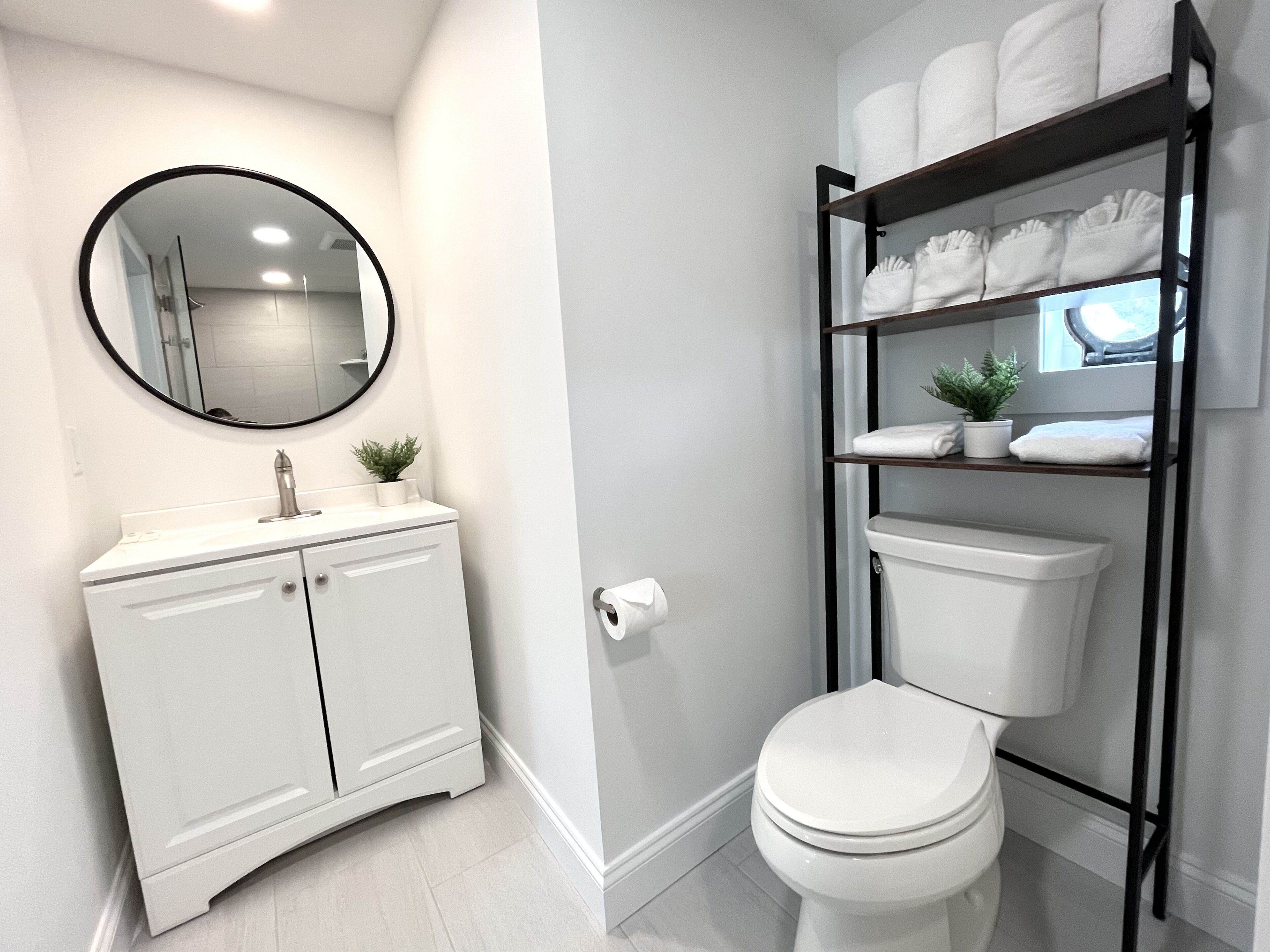 A modern bathroom with a white vanity, a round mirror, a toilet, and a shelf with towels and a plant.