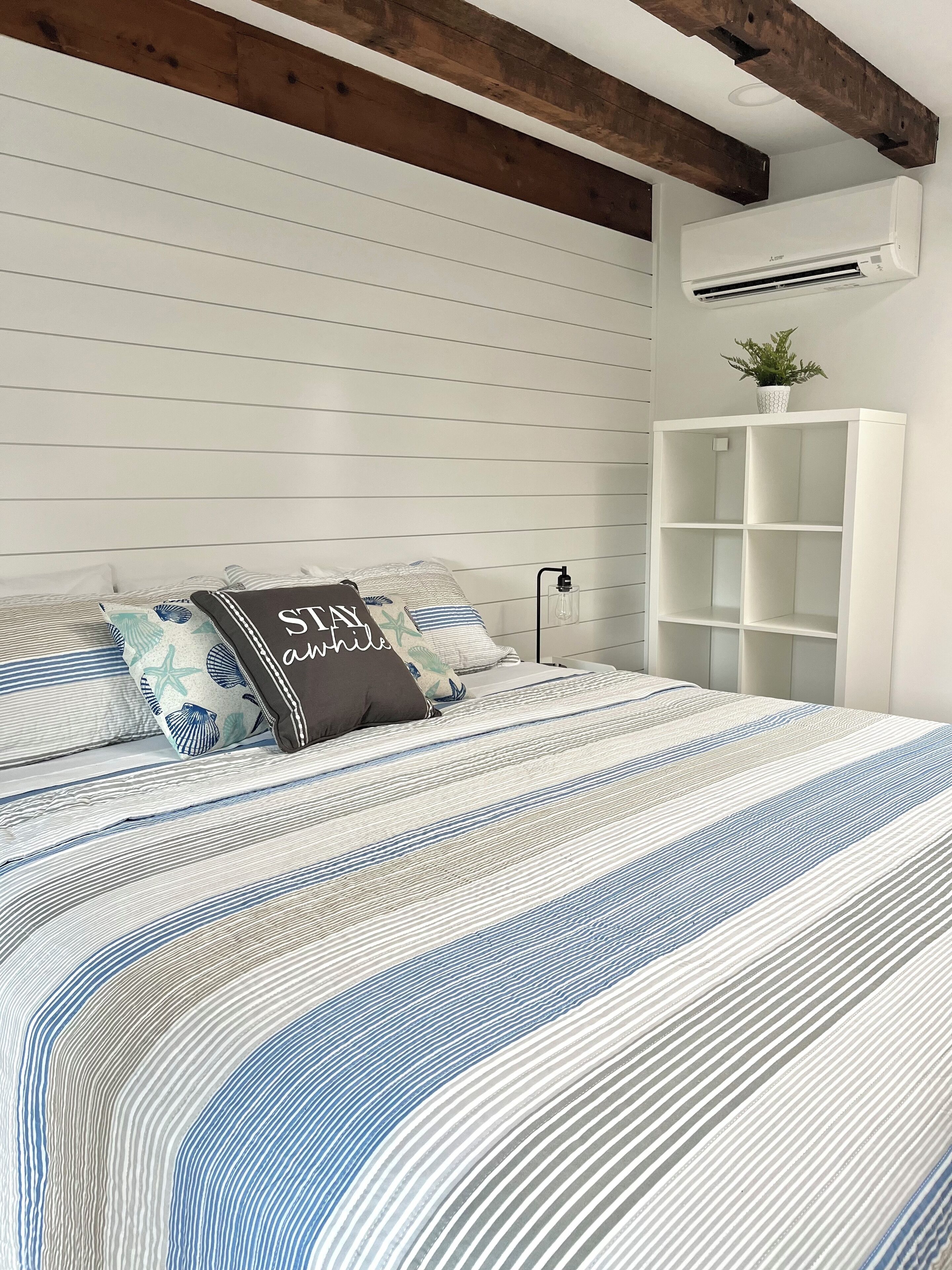 A bedroom with a striped bedspread, a white wall with horizontal lines, a wooden ceiling beam, a white air conditioning unit, and a white shelving unit.