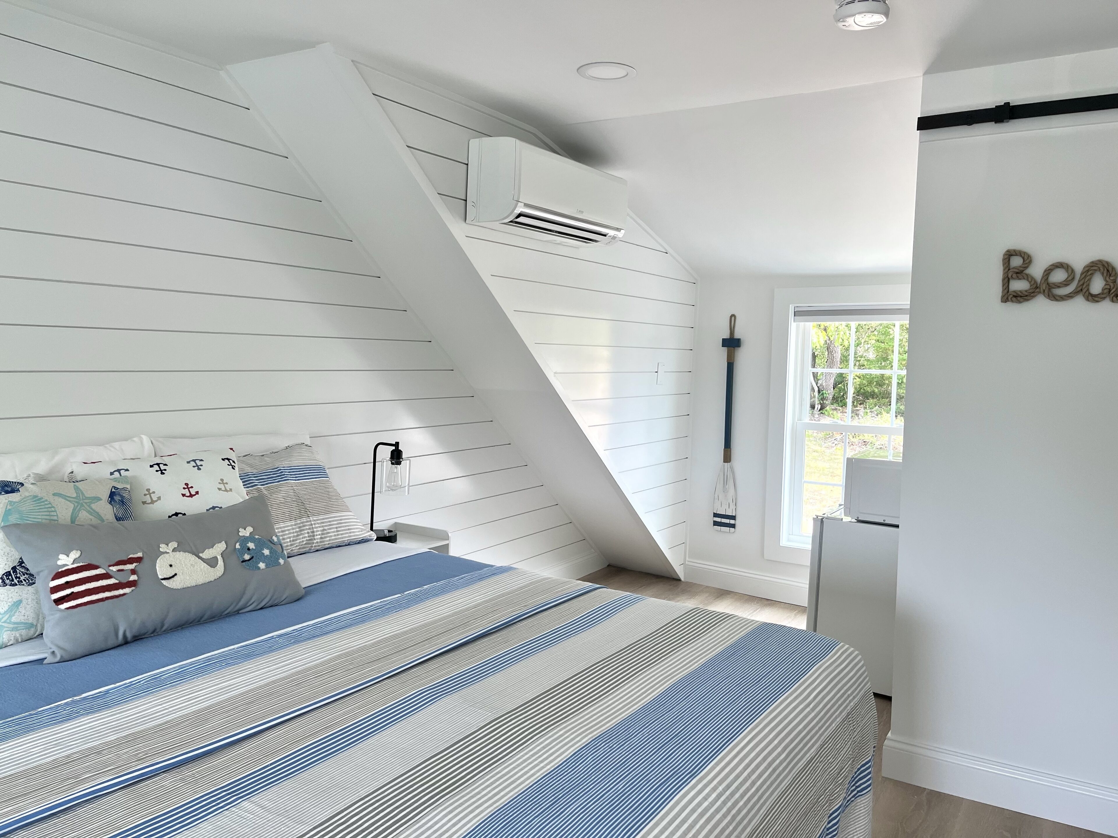 Un dormitorio con techo inclinado, una cama con ropa de cama a rayas, almohadas con temática náutica, una mesita de noche con lámpara y una tabla de remo fijada a la pared.