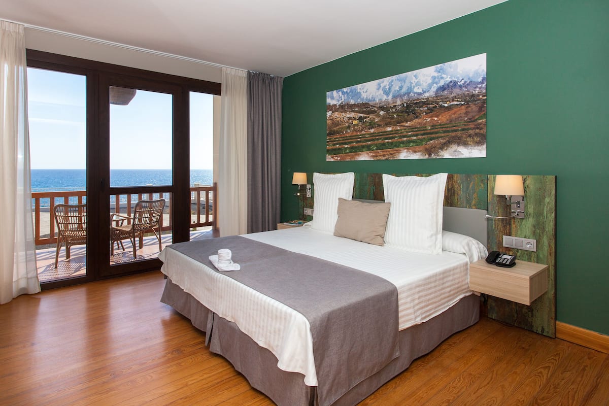 Une chambre d’hôtel avec un grand lit, une vue sur la mer, un tableau au mur et un balcon avec des chaises.