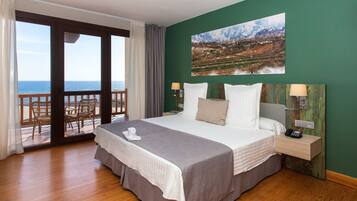 Une chambre d’hôtel avec un grand lit, une vue sur la mer, un tableau au mur et un balcon avec des chaises.