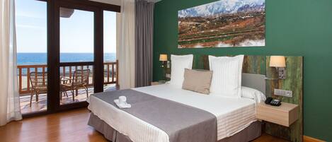 A hotel room with a large bed, a view of the sea, a painting on the wall, and a balcony with chairs.