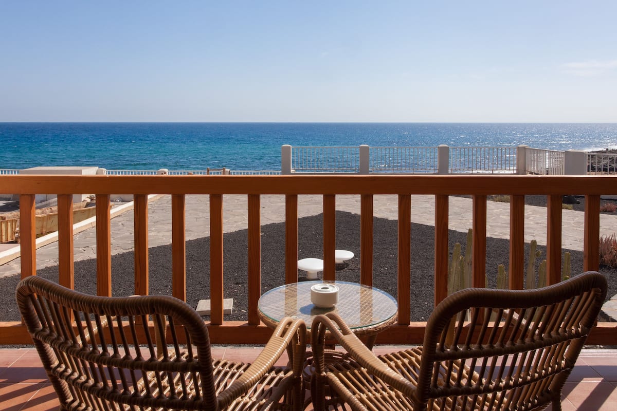 Un balcon avec des fauteuils en osier et une petite table ronde, donnant sur une plage et l’océan.