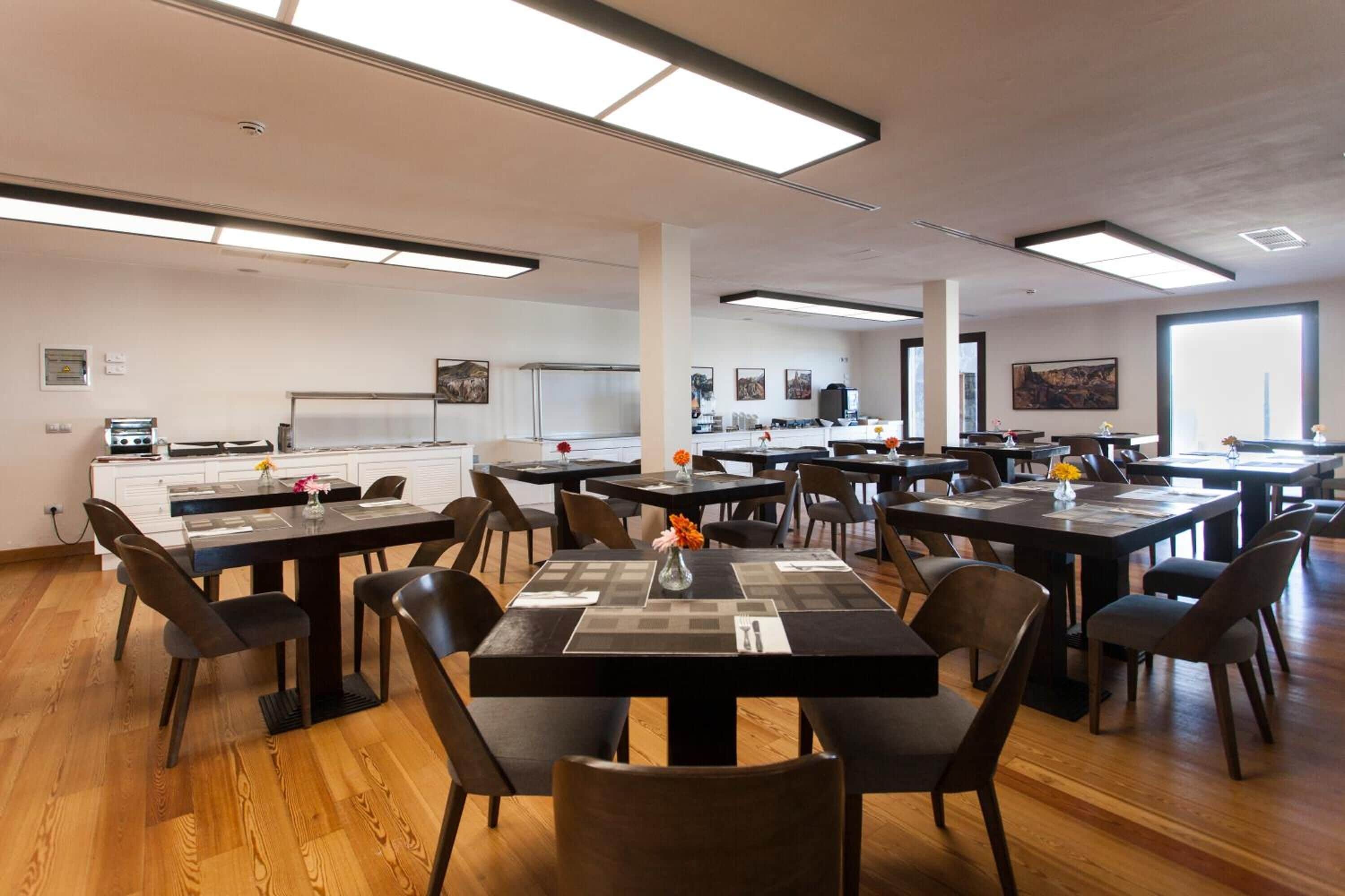 Un grand espace dédié à la salle à manger, avec plusieurs tables et chaises, un coin cuisine à l’arrière-plan et de grandes fenêtres qui laissent entrer la lumière naturelle.