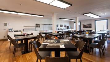 Un grand espace dédié à la salle à manger, avec plusieurs tables et chaises, un coin cuisine à l’arrière-plan et de grandes fenêtres qui laissent entrer la lumière naturelle.