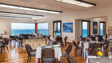 Un área de comedor con mesas dispuestas para los huéspedes, amplias ventanas con vista panorámica de la ciudad y piso de madera.