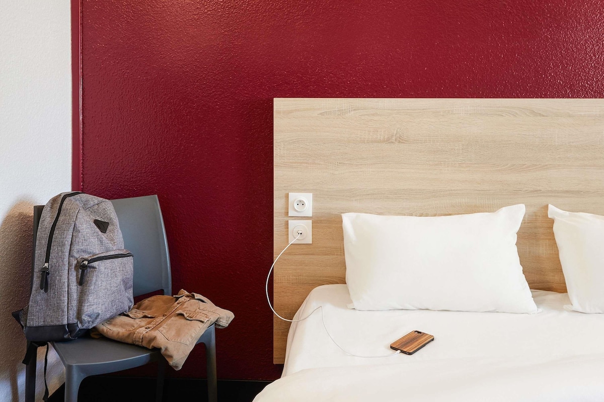 Una habitación de hotel con cama, una silla, una mochila y un enchufe en la pared.