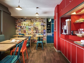 A café with wooden tables and colorful chairs, a red counter with a coffee machine, and a wall decorated with travel posters.