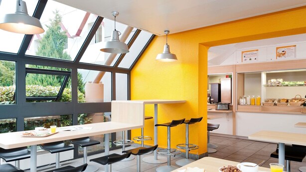 A modern cafe with a yellow accent wall, large windows, and a breakfast bar with seating. - hotelF1 Paris Gennevilliers (Gennevilliers)