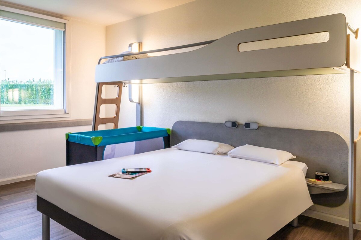 Uma cama beliche com escrivaninha e cadeira, uma mesa de cabeceira com abajur, e uma janela com vista para o verde.