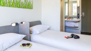 Een hotelkamer met een bed, kussens, een nachtkastje met een boek en hoofdtelefoon, een spiegel, en een badkamer met een wastafel en handdoekhouder.