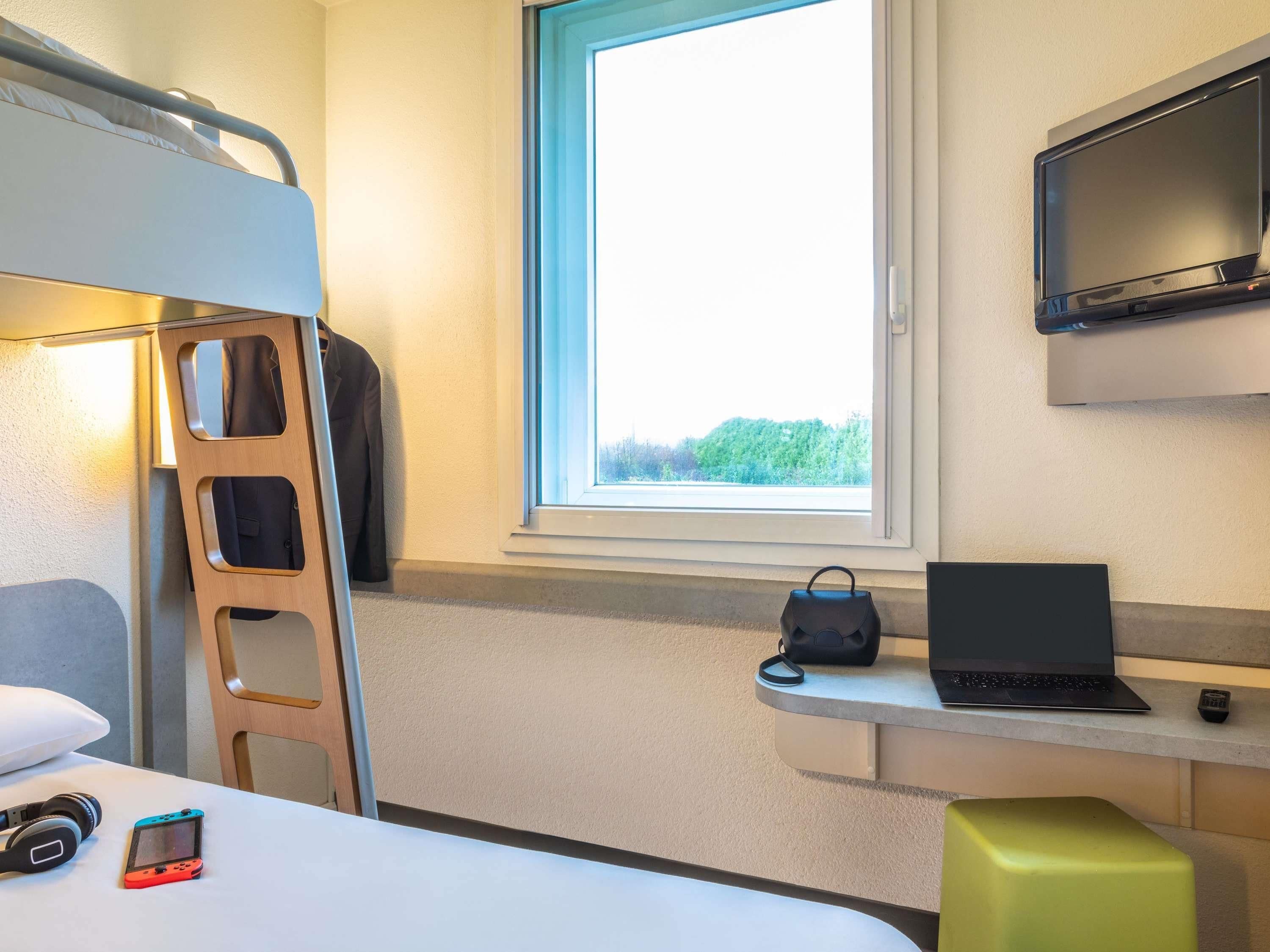 A compact hotel room with a bunk bed, a desk with a laptop, a TV, and a window with a view of trees.