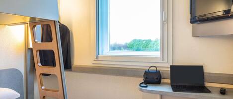 A compact hotel room with a bunk bed, a desk with a laptop, a TV, and a window with a view of trees.
