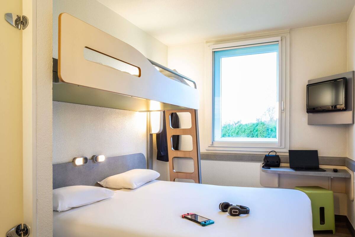 Une chambre d’hôtel moderne équipée d’un lit superposé, d’un bureau avec un ordinateur portable, d’une télévision et d’une fenêtre donnant sur un paysage verdoyant.