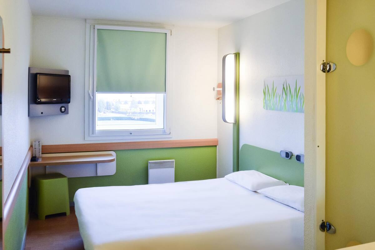 Una habitación de hotel con cama, televisión, ventana, mesita de noche y baño.
