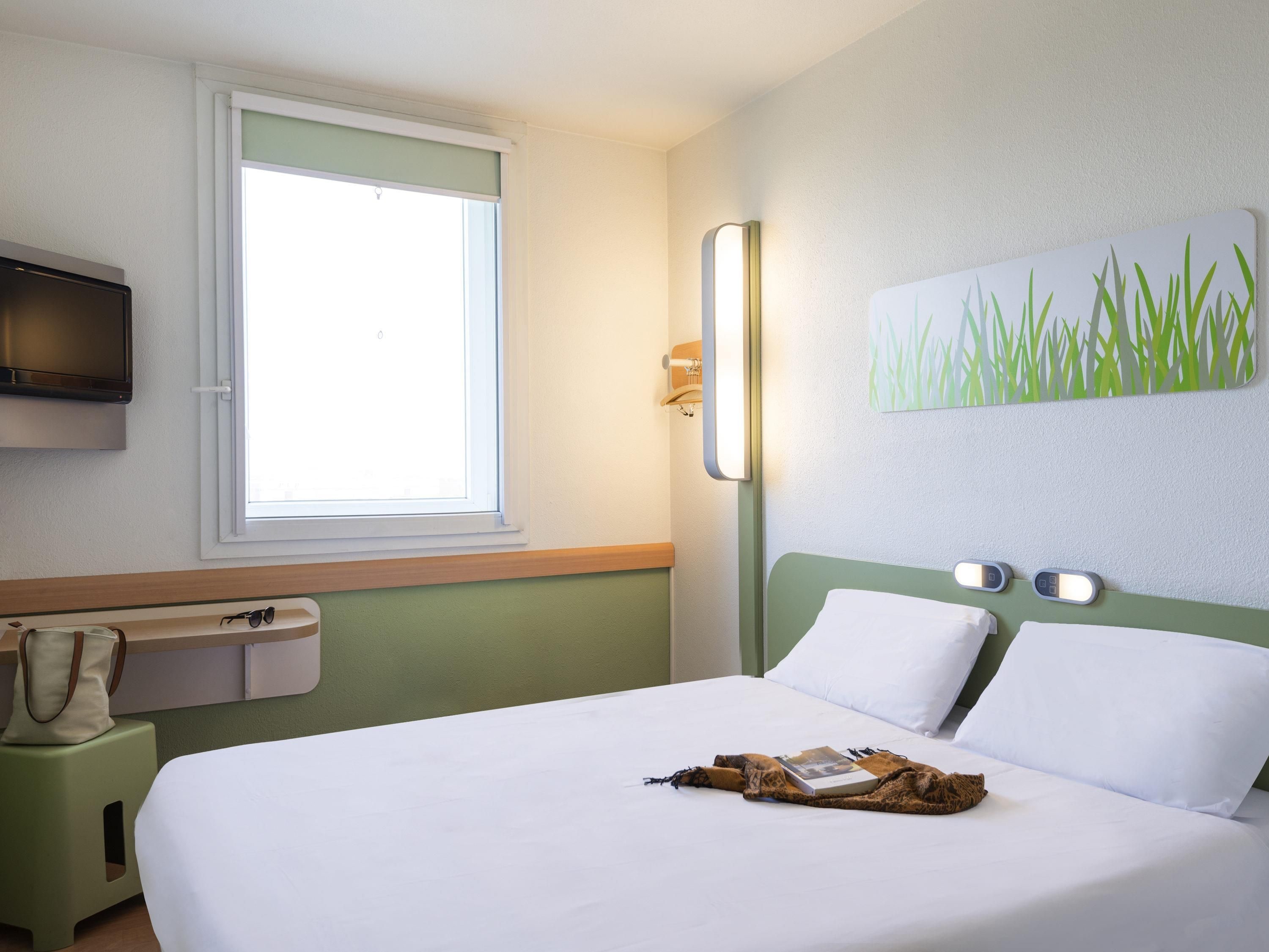 Quarto de hotel com uma cama, janela, televisão, mesa de cabeceira e luz acoplada à parede.