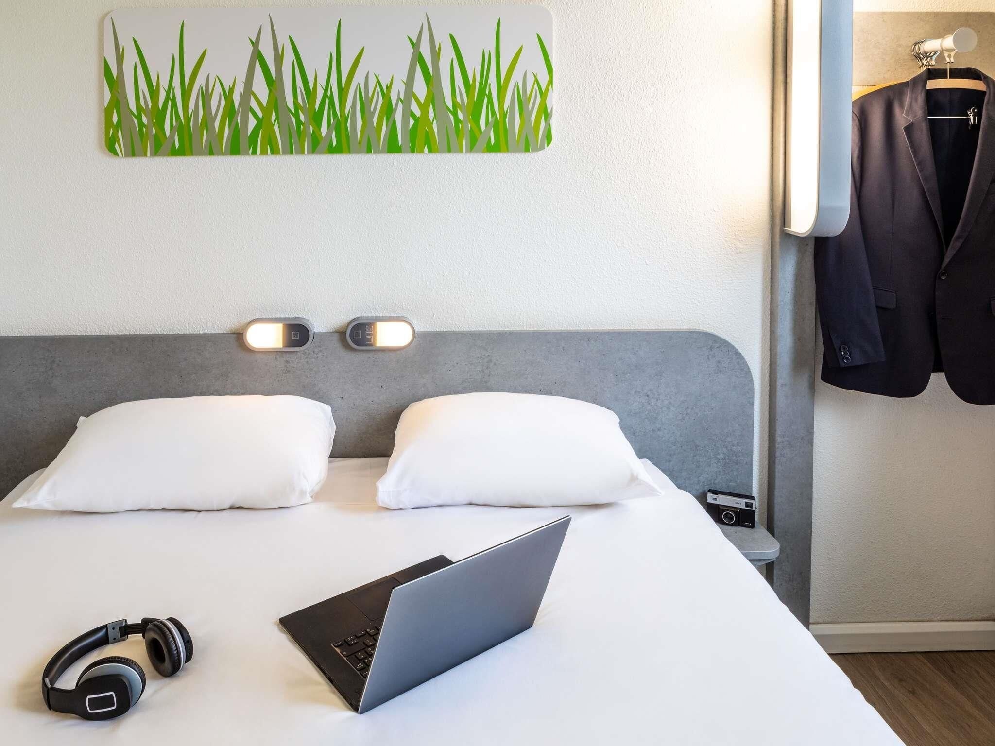 Habitación de hotel con cama, almohadas, una computadora portátil, audífonos y una lámpara fijada en la pared.