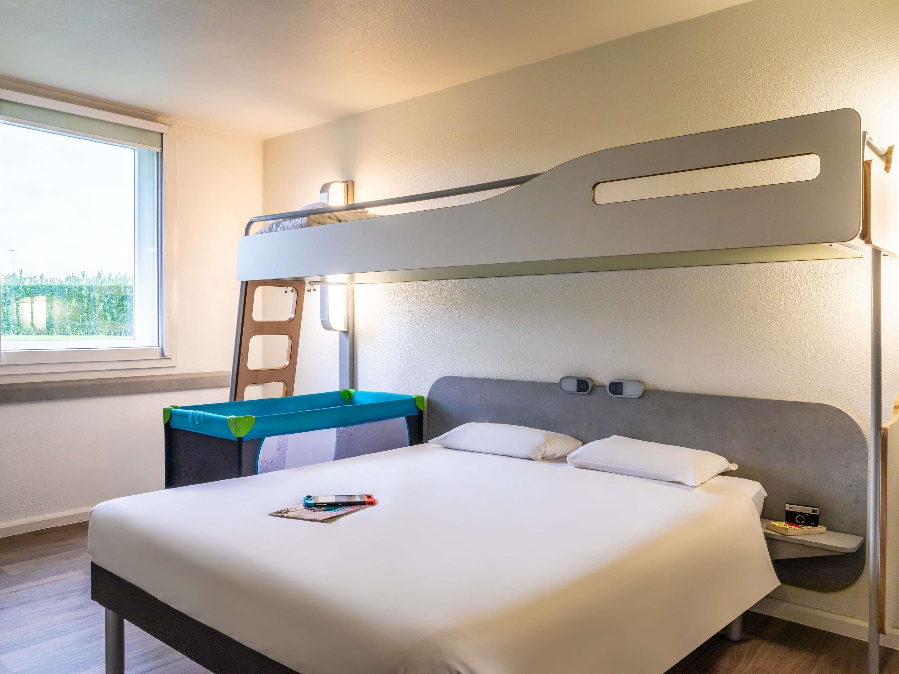 A bunk bed with a desk and chair, a bedside table with a lamp, and a window with a view of greenery.