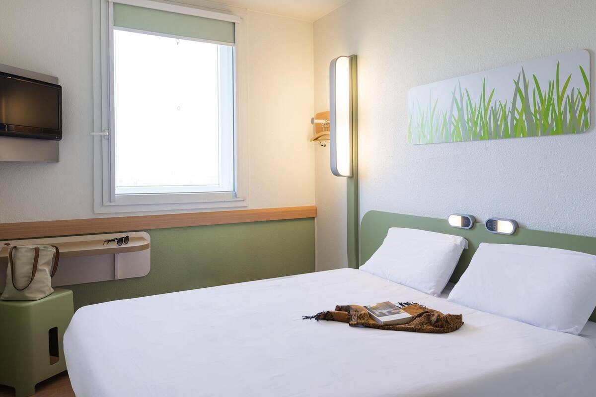 Une chambre d’hôtel équipée d’un lit, d’une fenêtre, d’une télévision, d’une table de chevet et d’une lampe fixée au mur.