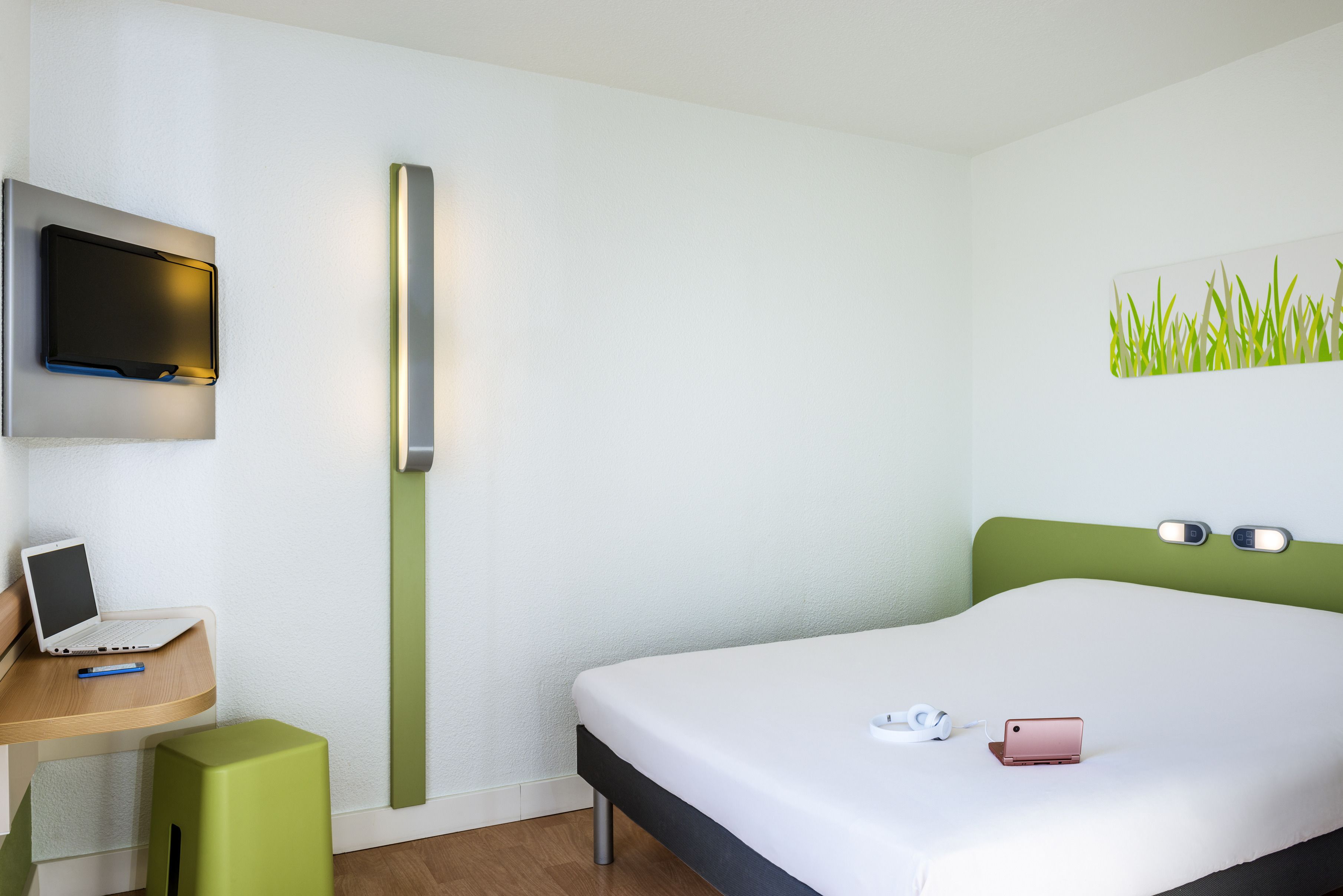 Une chambre d’hôtel moderne équipée d’un lit, d’un bureau avec un ordinateur portable, d’un téléviseur fixé au mur et d’une table de chevet avec un téléphone et des écouteurs.