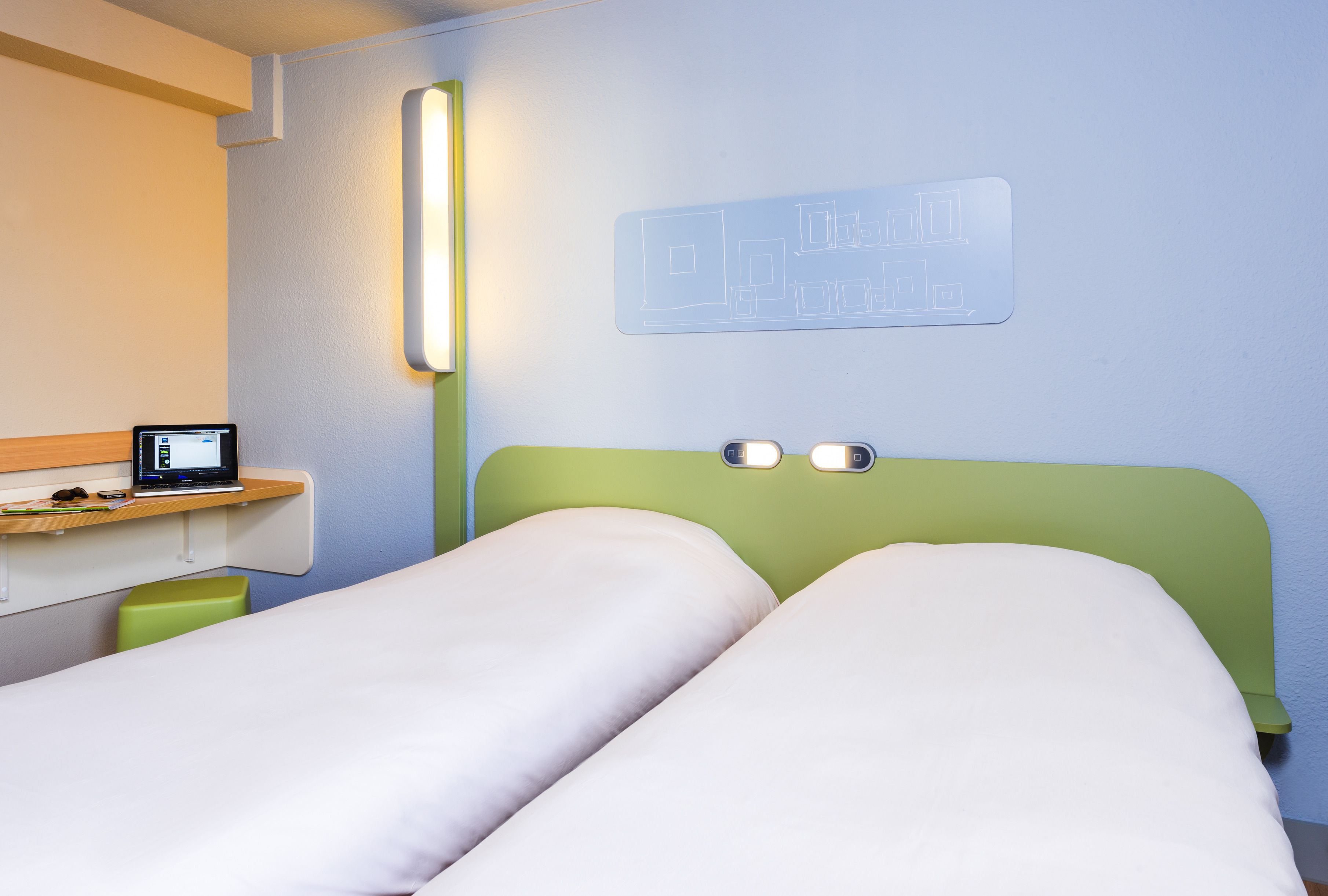 A hotel room with two beds, a desk with a computer, and a wall-mounted light fixture.