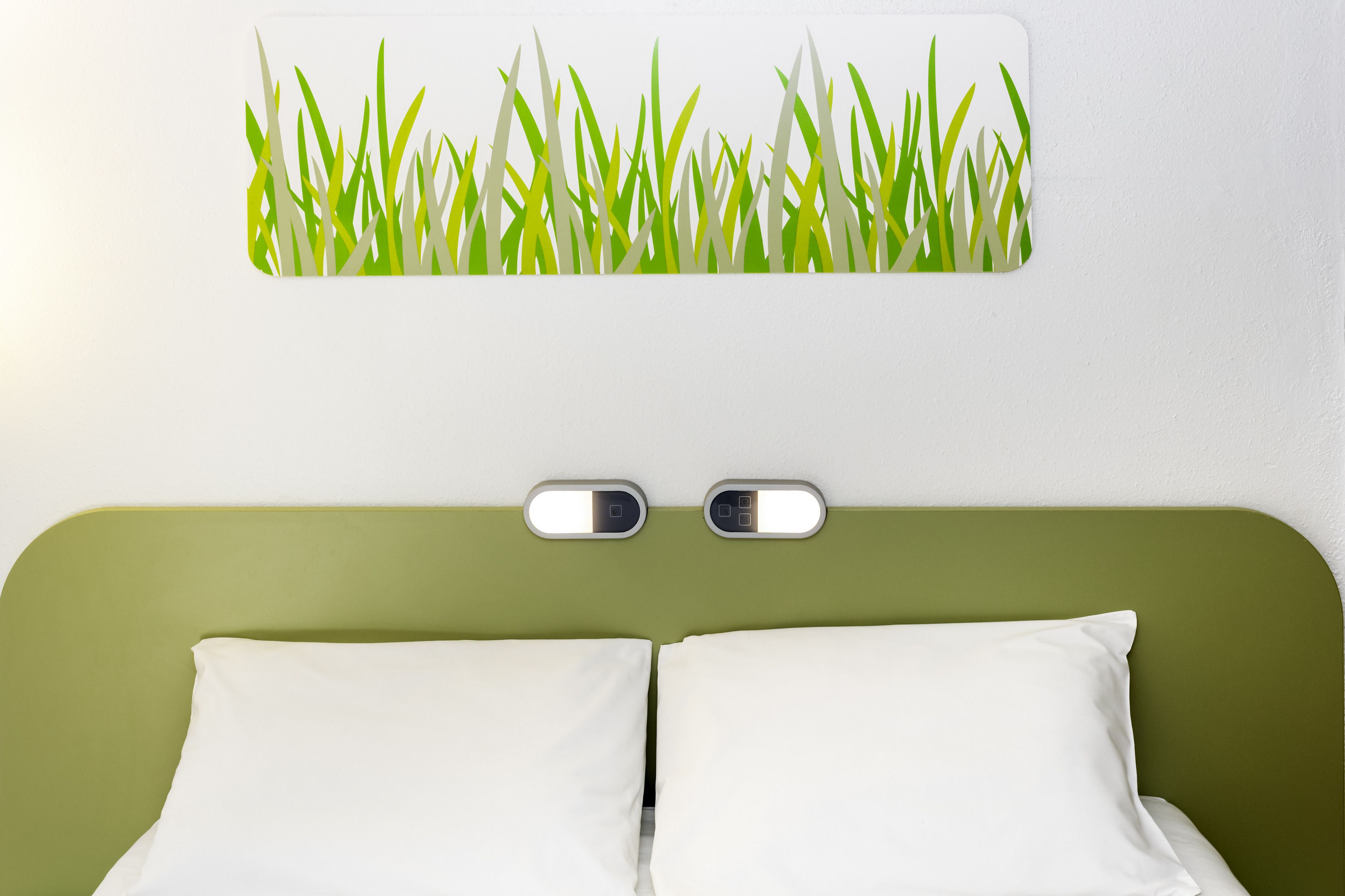 A hotel room with a green headboard, two white pillows, and a wall-mounted light fixture.