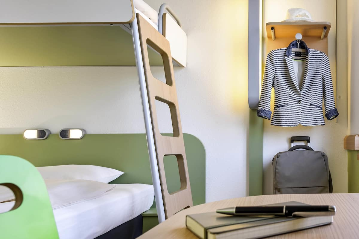 Un lit superposé avec une tête de lit verte, une table de chevet avec un livre et un stylo, une valise et un porte-manteau mural avec une veste à rayures.