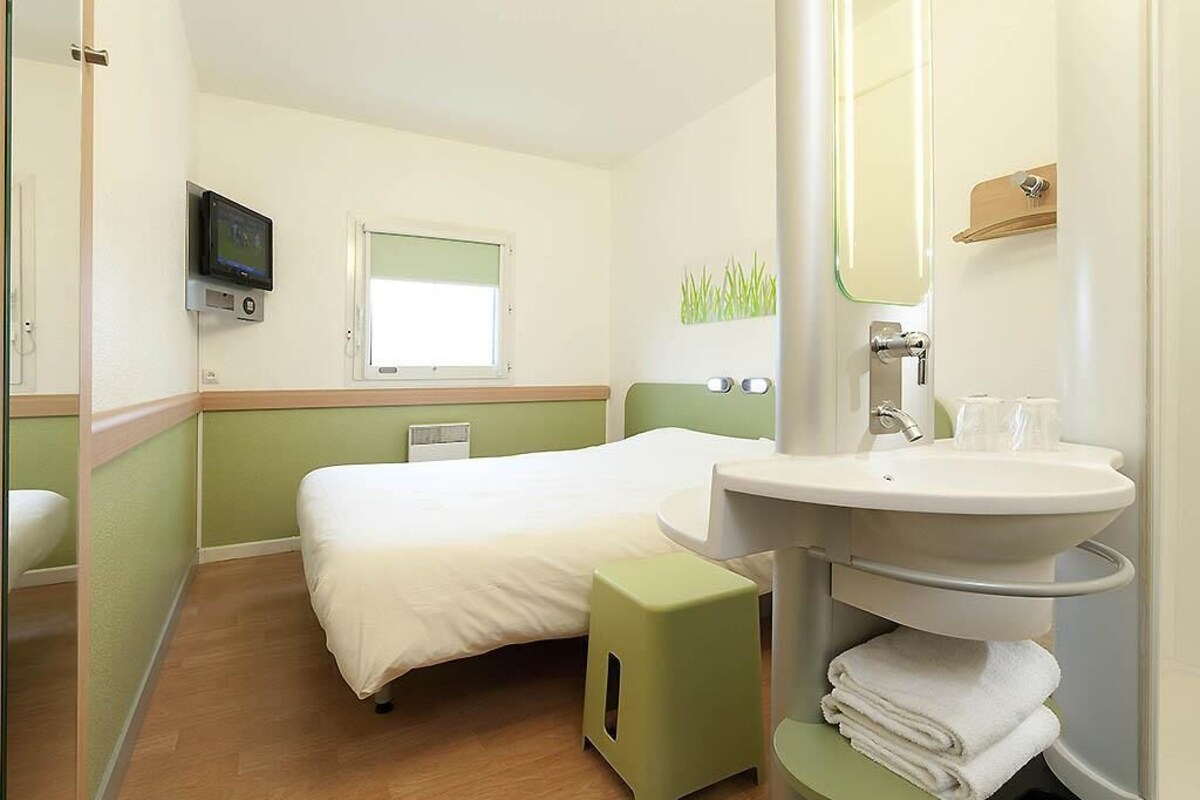 Ein Hotelzimmer mit einem Bett, einem grünen Hocker, einem Waschbecken mit Spiegel und einem Fernseher an der Wand.