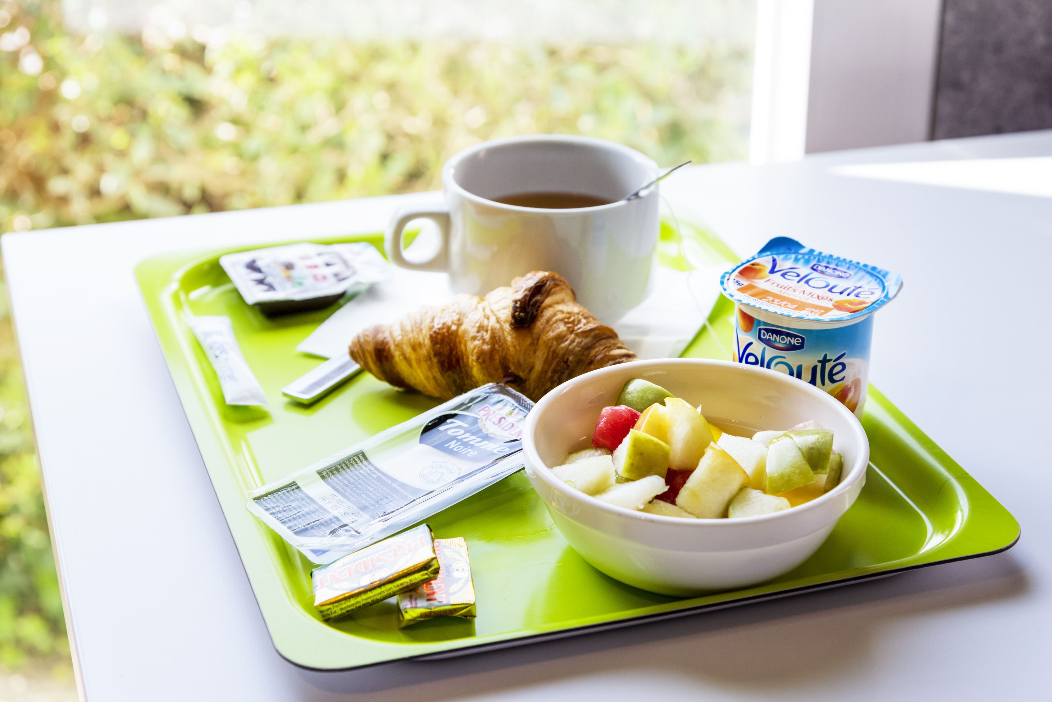 A breakfast tray with a croissant, a cup of coffee, a bowl of fruit, and a container of yogurt.