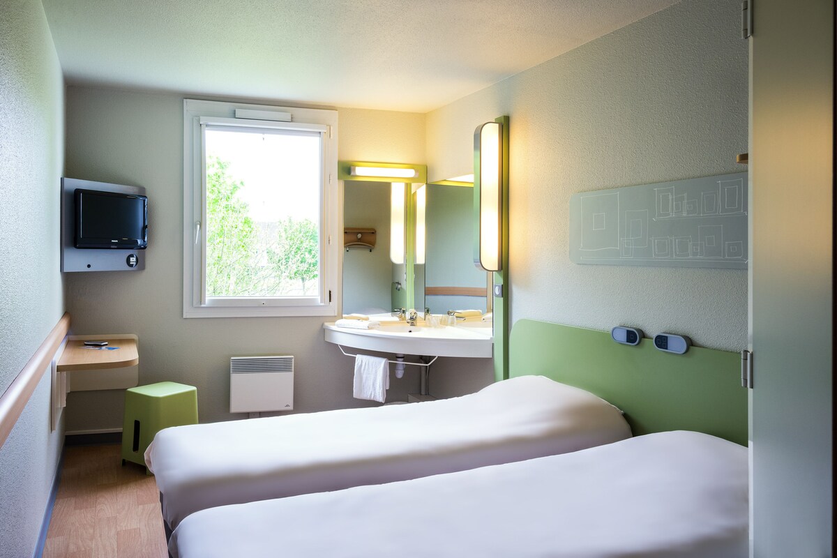 Ein Hotelzimmer mit zwei Betten, einem Waschbecken, einem Spiegel, einem Fernseher und einem Fenster.