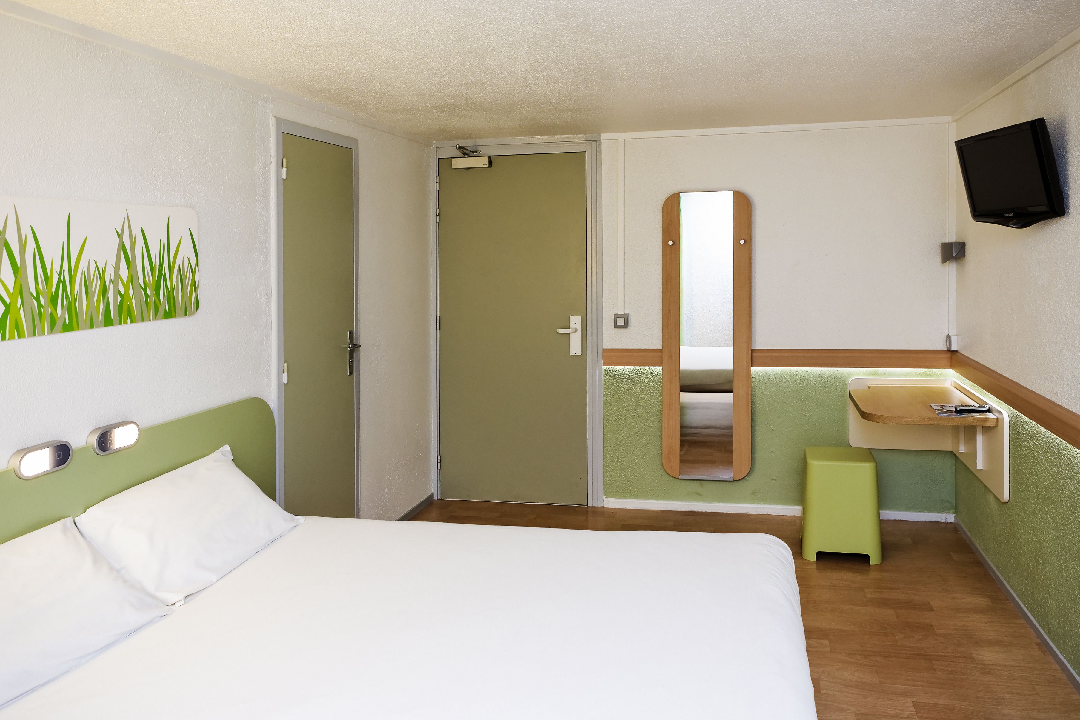A hotel room with a bed, a green wall, a mirror, a TV, and a small stool.