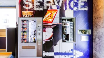 Un distributeur automatique, équipé d’un appareil à boissons et d’une machine à café, est installé dans un hall d’entrée moderne, au-dessus duquel on peut lire un grand panneau indiquant « SERVICE ».