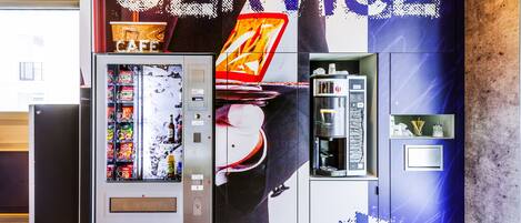 A vending machine with a beverage dispenser and a coffee machine, located in a modern lobby with a large 'SERVICE' sign above.