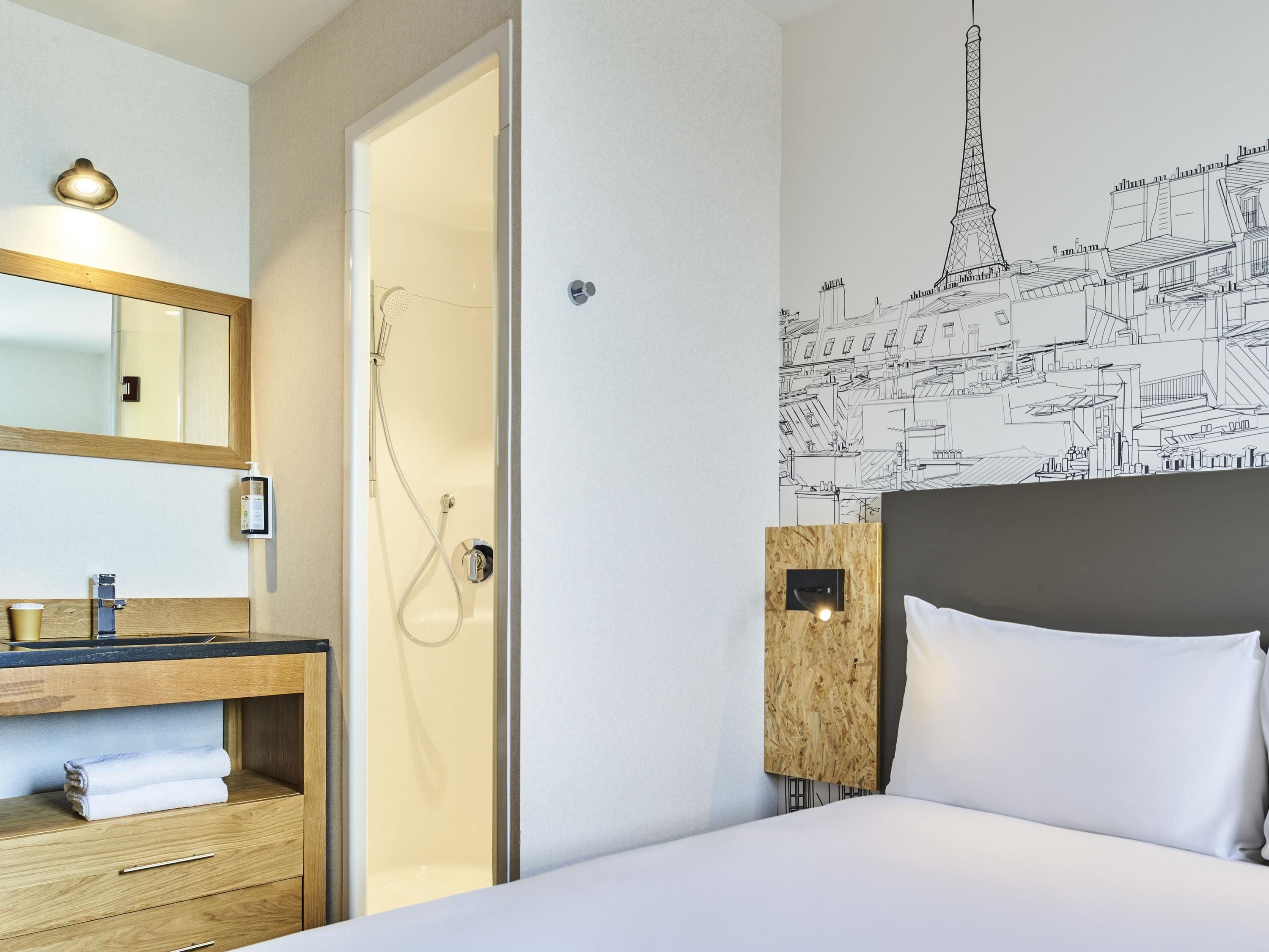 A hotel room with a wooden vanity, a shower, a bed with white bedding, and a wall mural of a cityscape.