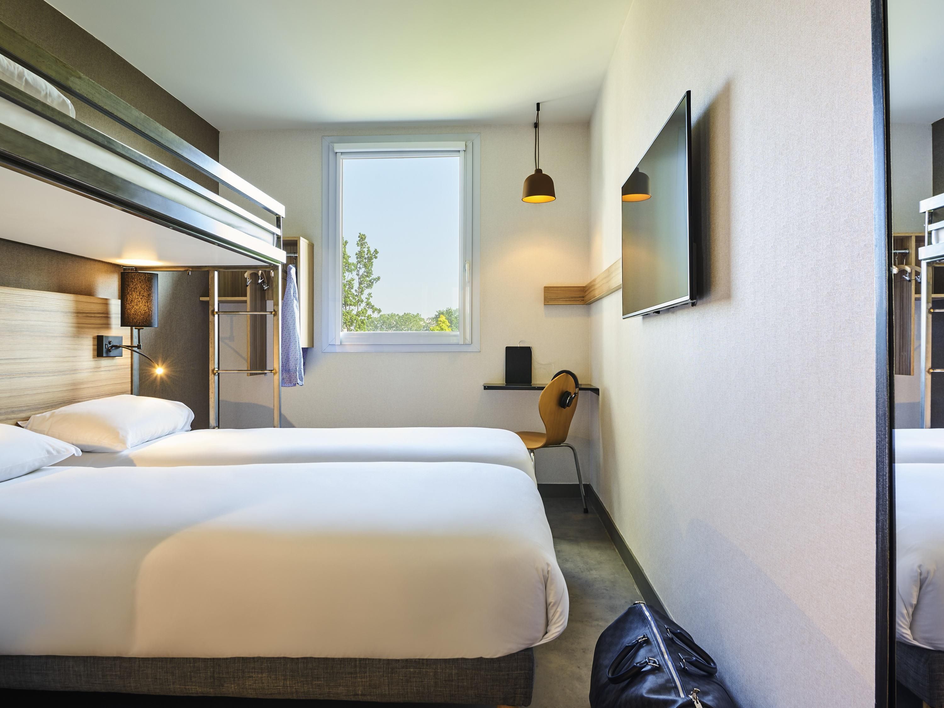 Une chambre d’hôtel avec deux lits, un bureau, une chaise, une télévision et une fenêtre donnant sur des arbres.