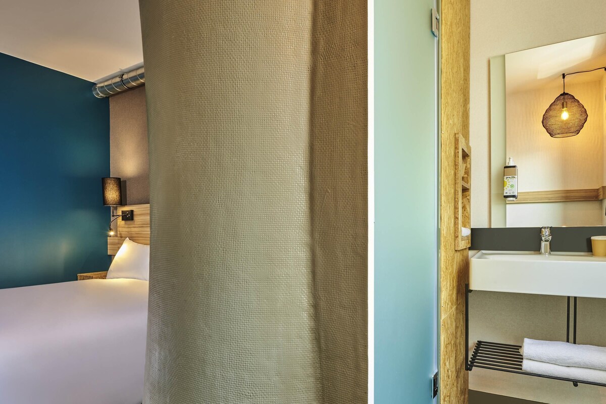Habitación de hotel con cama, lámpara de pared, espejo, lavabo y toallero.