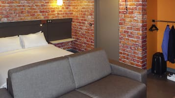 Ein Hotelzimmer mit Backsteinwand, einem Bett, einem grauen Sofa, einem Kleiderschrank und einem Kleiderhaken.