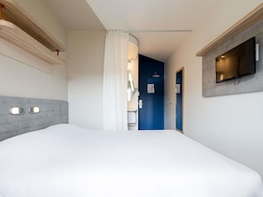 Een hotelkamer met een bed, een aan de muur bevestigde televisie, een deur naar een badkamer en een houten plank boven het bed.