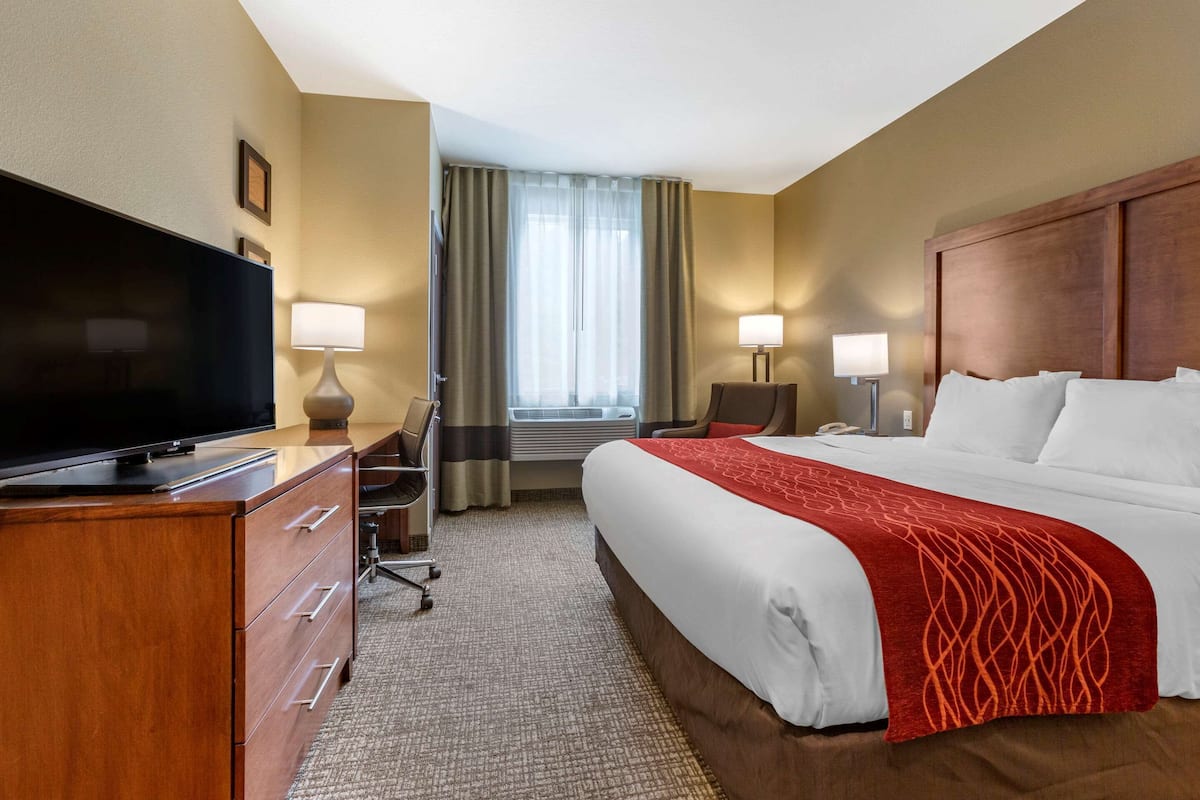 Une chambre d’hôtel dotée d’un grand téléviseur à écran plat, d’une commode en bois, d’un lit avec du linge de lit blanc et une couverture rouge, de deux lampes de chevet et d’une fenêtre avec des rideaux.