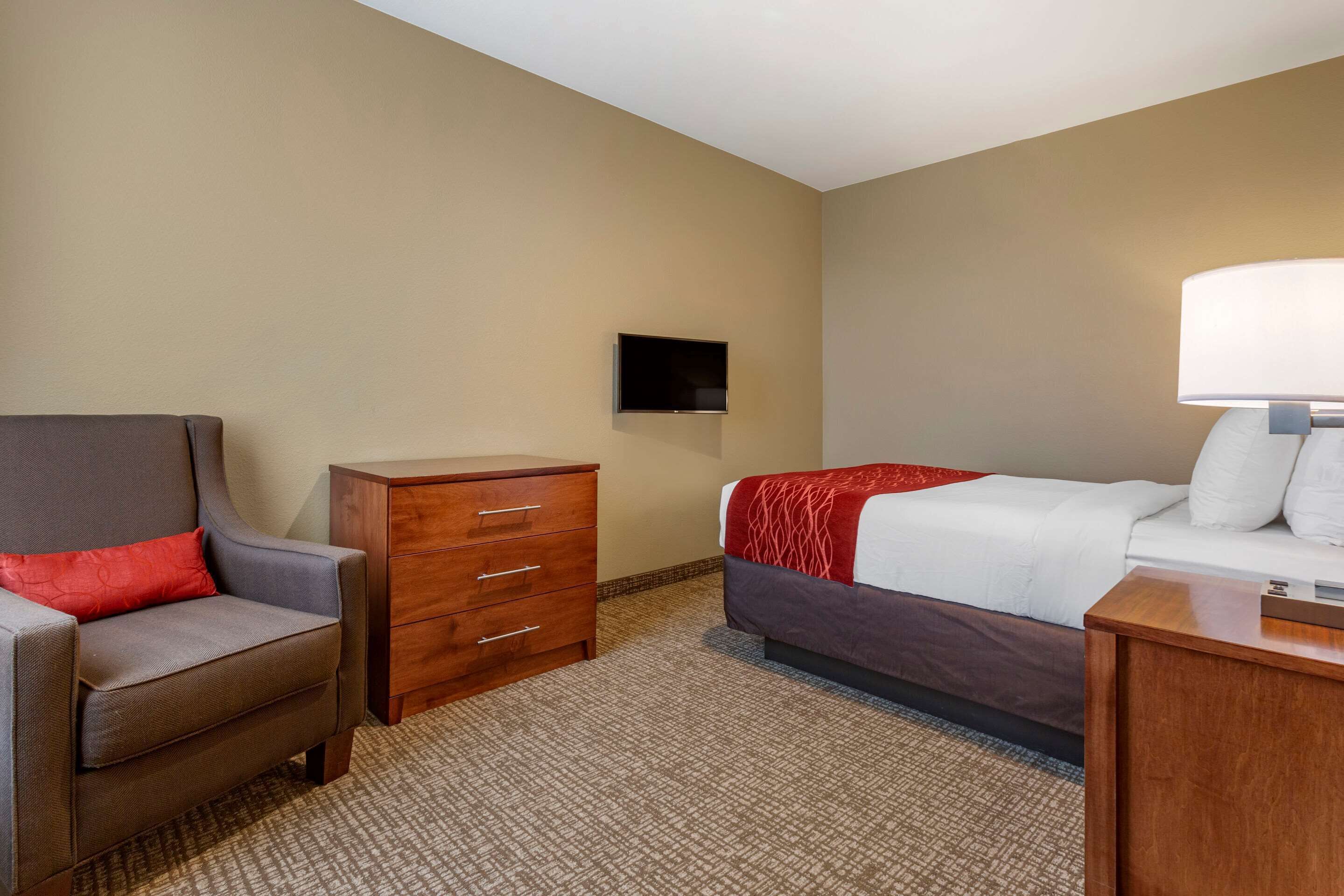 Quarto de hotel com cama, cadeira, cômoda e TV fixada na parede.