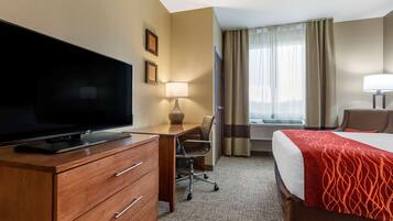 Une chambre d’hôtel avec un téléviseur à écran plat posé sur une commode en bois, un bureau avec une chaise, un lit recouvert d’une couverture rouge et une fenêtre avec des rideaux.