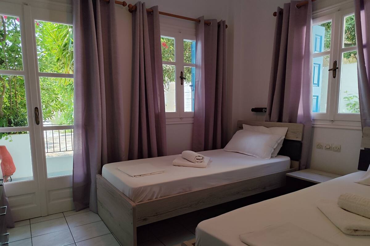 A bedroom with a bed, a bedside table, a window with curtains, and a view of greenery outside.