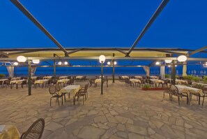 An outdoor dining area with tables and chairs, a stone floor, and a view of the sea at dusk. - Olympios Zeus (Dio-Olympos)