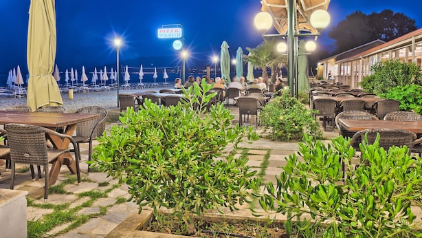 An outdoor seating area with tables and chairs, a sign for 'PIZZA SPAGHETTI', and a view of the beach at night.