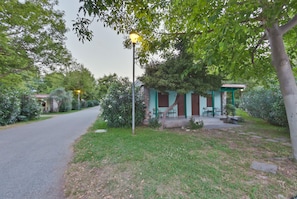 A small, single-story building with a porch and outdoor seating area, surrounded by greenery and a paved path.