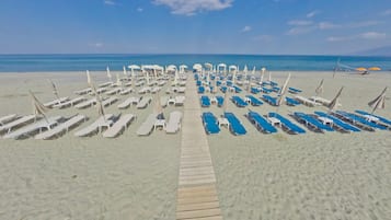 A beach with rows of sun loungers and umbrellas, a wooden walkway leading to the water, and a clear blue sky.