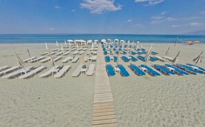 A beach with rows of sun loungers and umbrellas, a wooden walkway leading to the water, and a clear blue sky. - Olympios Zeus (Dio-Olympos)