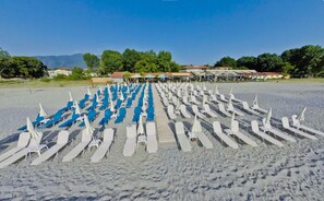 A beach with rows of sun loungers and umbrellas, a covered area with tables and chairs, and buildings in the background. - Olympios Zeus (Dio-Olympos)