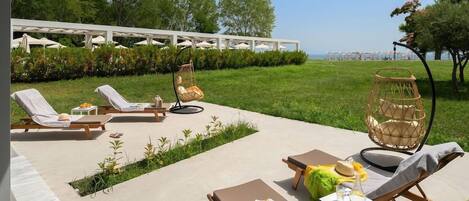 A sunlit outdoor seating area with lounge chairs and a small table, surrounded by greenery and a view of the sea.